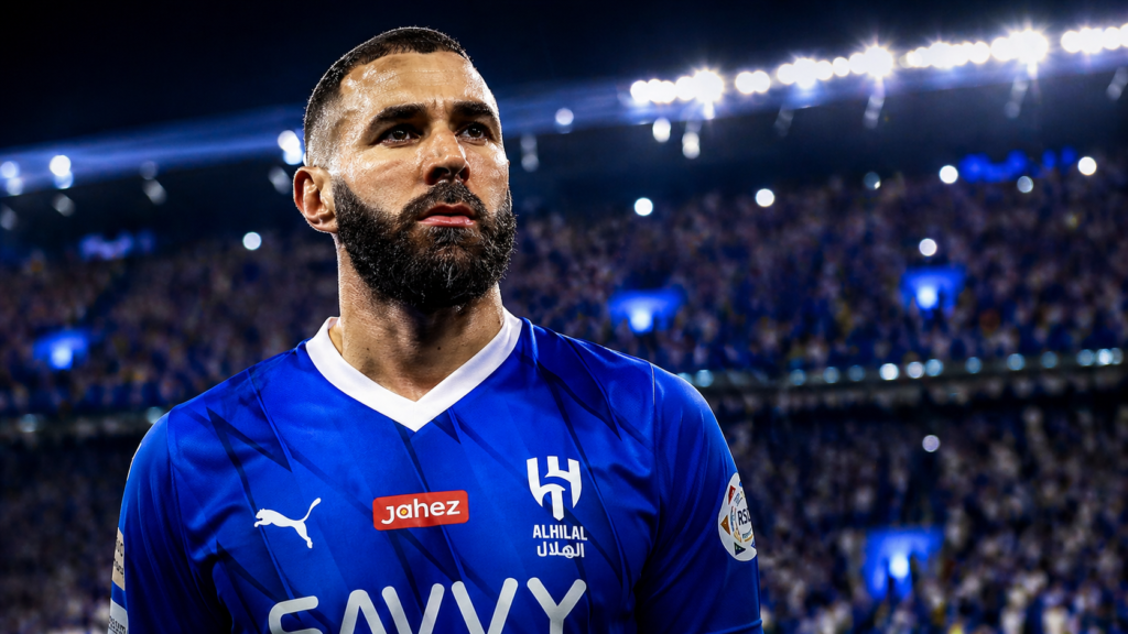 A highly detailed, cinematic sports portrait of a male footballer resembling Karim Benzema standing in a packed stadium at night. He wears a blue Al Hilal-style jersey, with visible sweat on his face and a focused, confident expression. Bright stadium floodlights glow behind him, while the crowd is blurred to create a shallow depth-of-field effect. The image has high contrast, dramatic lighting, and a wide 16:9 aspect ratio, giving it an inspirational and legendary atmosphere.